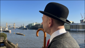 head shot - Scott Rochester is standing on London Bridge looking away from the photographer towards Tower Bridge - he is wearing a jacket and tie, and a bowler hat - he is carrying an umbrella on a bright sunny day without a cloud in the sky (this is his Patrick Macnee impression) - in the middle distance is the River Thames - beyond that the buildings of the South Bank including the London Bridge Hospital and the Hays Galleria are visible - a distant Tower Bridge is visible on the left hand side - slightly to the right and in front of that is HMS Belfast