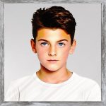 A watercolour painting of a 13 year old boy- he has neat dark brown hair, trimmed nicely at the sides and plentiful on top with a faint wave. His eyes are a beautiful blue. His face has a neutral expression with the merest hint of a smile. He is wearing a white T shirt against a plain grey background. The image is labelled AngryBard. He’s never angry unless you interfere with his book reading habit.