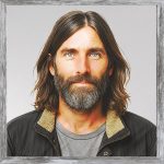 A watercolour painting of a 40 year old hobo with long, dark hair, a beard, and a scruffy appearance. A wandering story teller, he sleeps in the woods at night. He is wearing a grubby wolfskin jacket over an old grey T shirt. In spite of his rough looks and shabby clothes Benet is one of Wolfland's nicest characters.